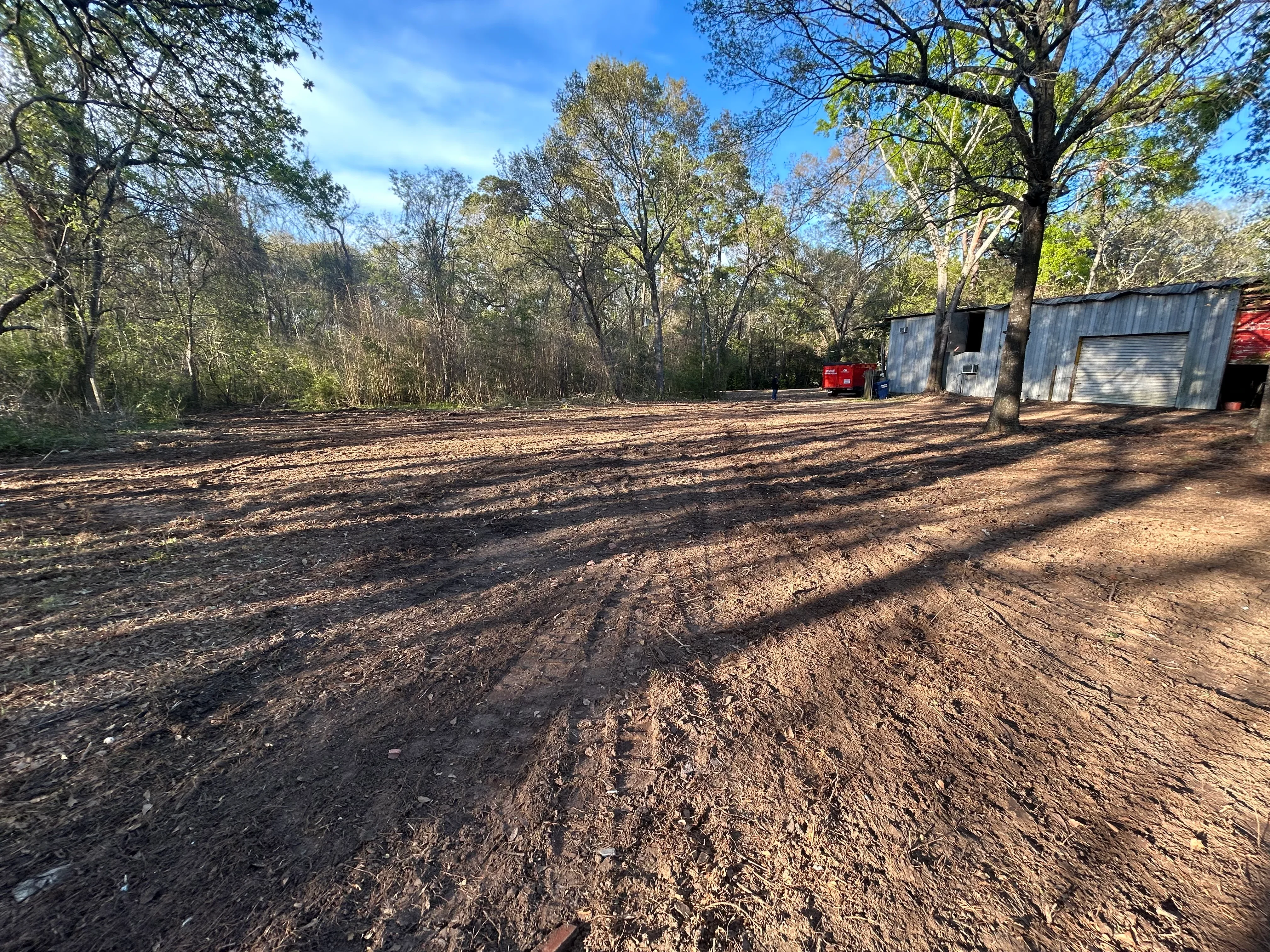 Cleared dirt lot with mature trees, metal storage structures, and brush removal completed