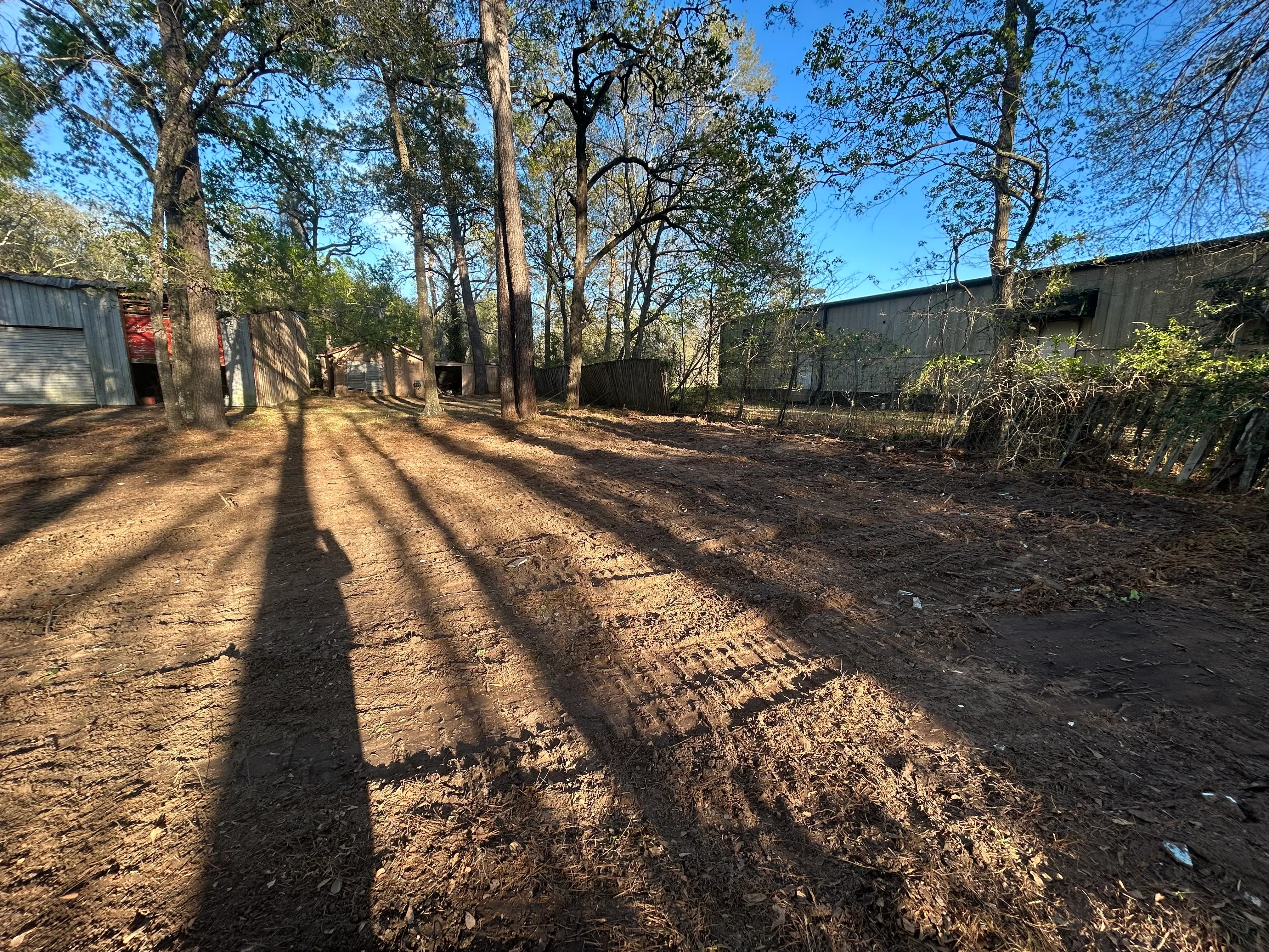 Cleared dirt lot with tall trees, graded soil, and metal structures in background