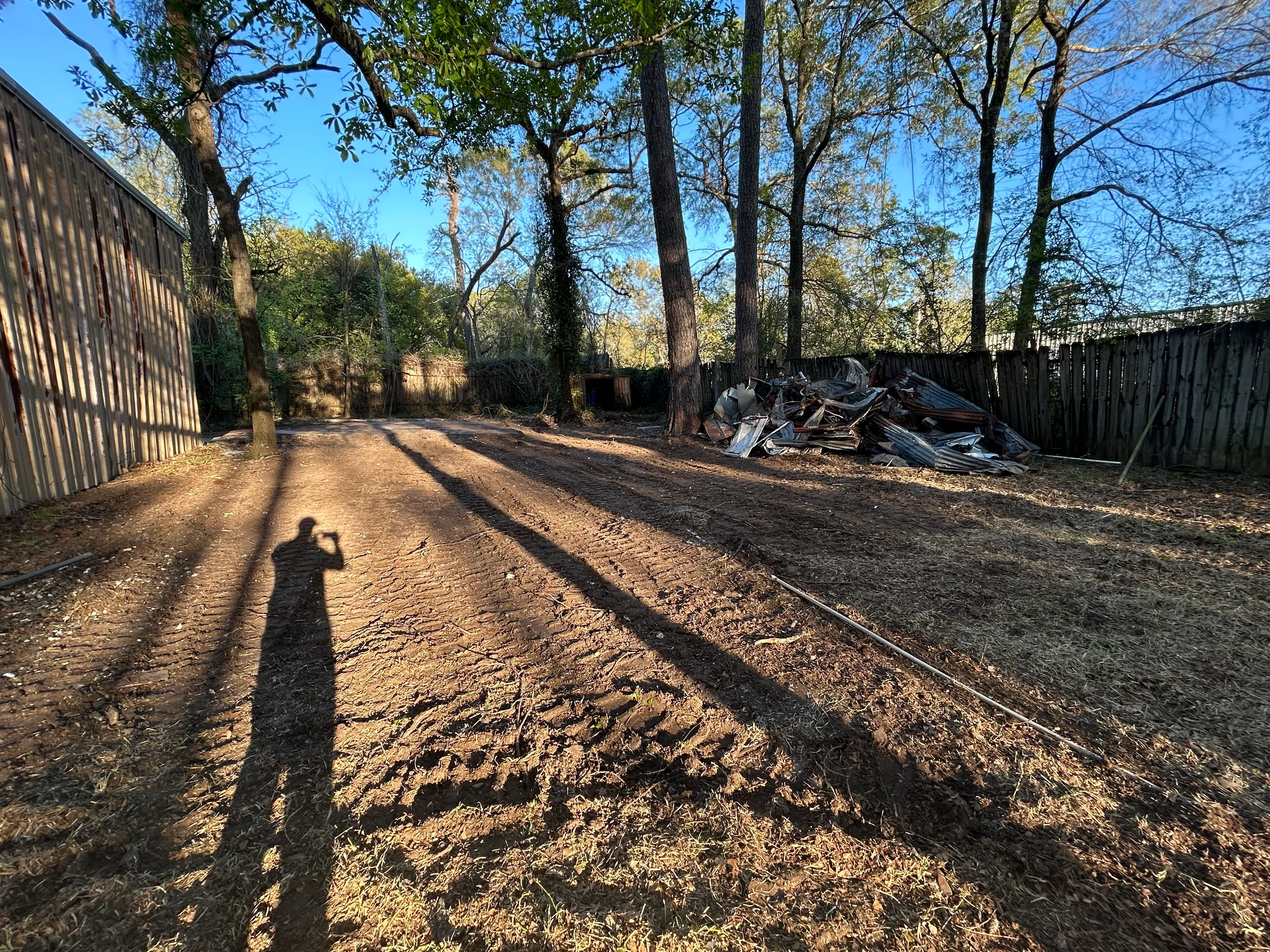 Cleared residential lot with gravel base, debris pile, wooden fence, and mature trees