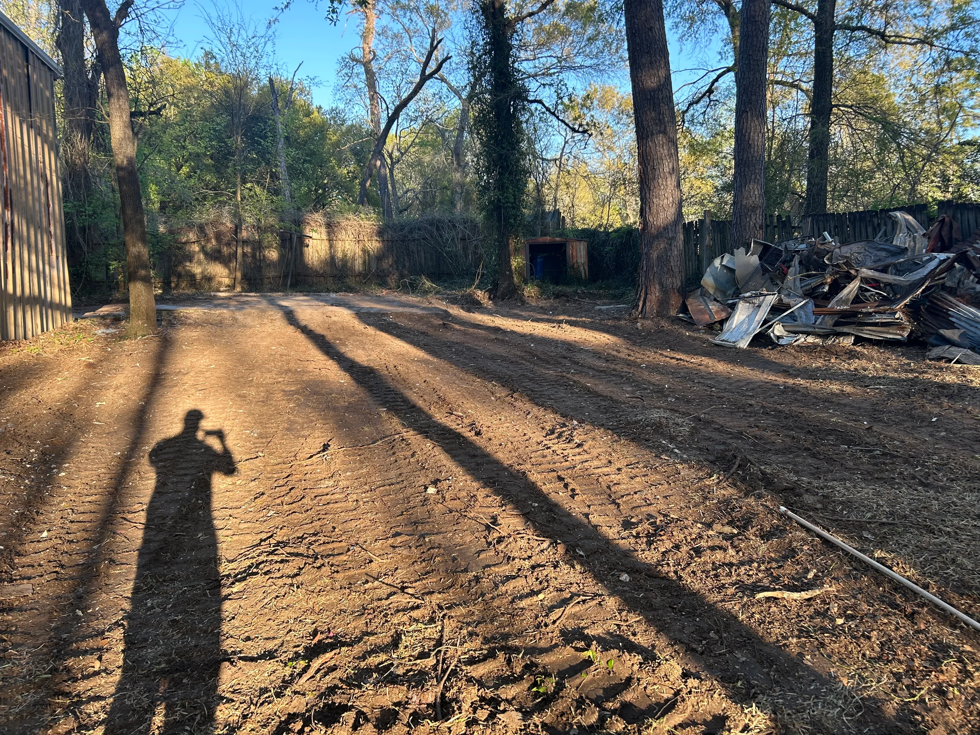 Cleared dirt lot with mature trees, metal debris pile, and metal fencing on wooded property